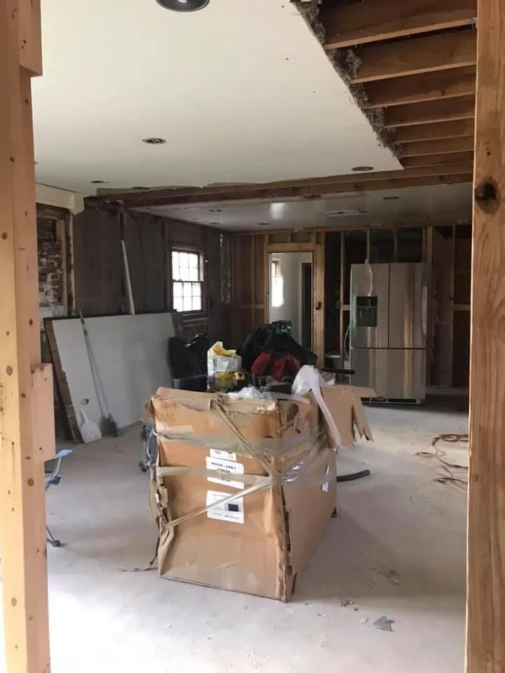 Interior of a house under renovation with exposed framework and construction tools, including a large damaged cardboard box in the center.