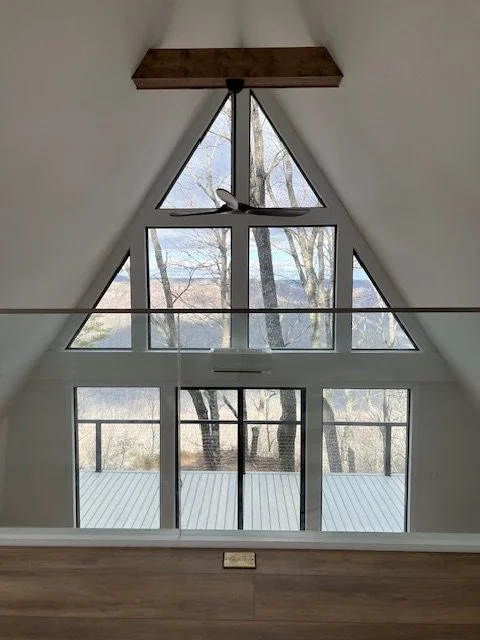 An interior view of an A-frame house with large triangular and rectangular windows, showing trees and a body of water outside. A ceiling fan is mounted near the apex of the windows.