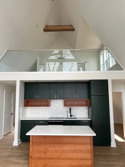 Modern kitchen with dark green cabinets, a white marble countertop island, and black appliances. Wooden accents and a high vaulted ceiling with a glass railing overlooking the space.