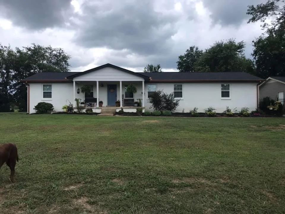 A single-story white house with a dark roof, front porch with steps, and potted plants. The yard has green grass and is partly cloudy.