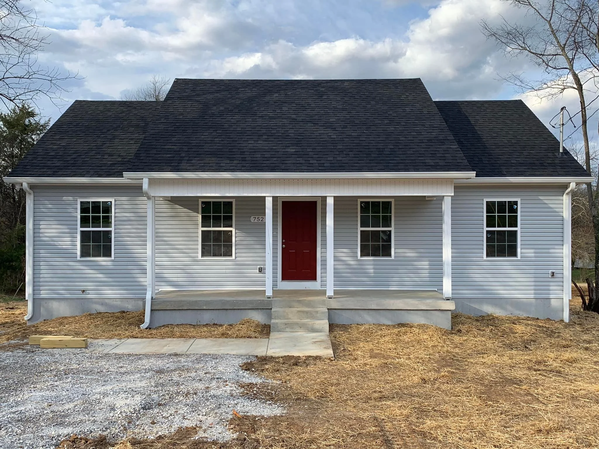 A newly constructed house with light gray vinyl siding, a dark shingled roof, four windows, and a red front door. There is a small concrete porch with steps, a gravel driveway, and patches of dirt and straw in the yard.