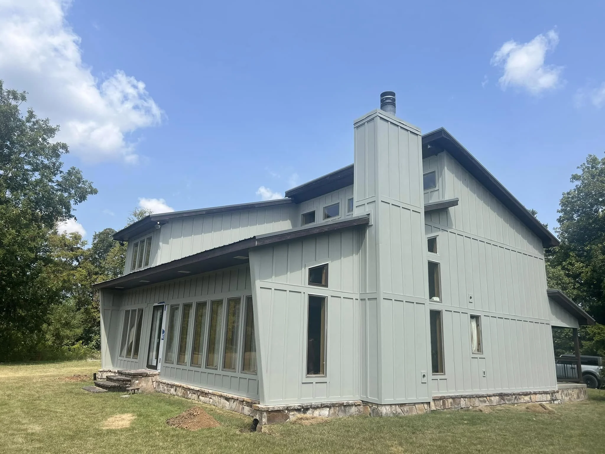 Modern light gray house with multiple windows, a stone foundation, and a chimney, surrounded by green grass and trees under a blue sky with some clouds.