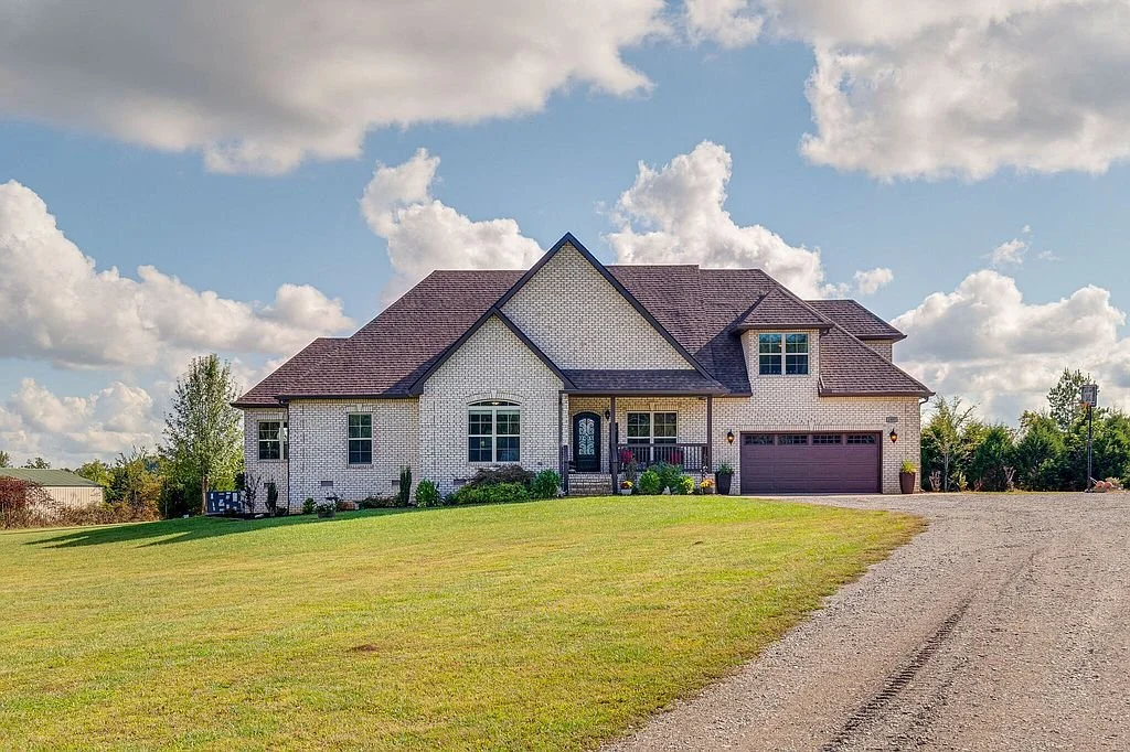 A large suburban house with a brick exterior, brown roof, and attached two-car garage, sitting on a spacious grassy lawn under partly cloudy sky.