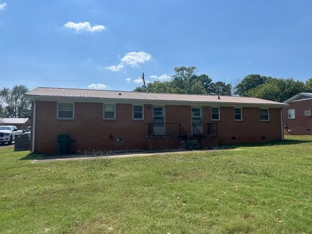 A single-story brick building with multiple windows and a small staircase leading to a front door, set in a grassy area under a blue sky with a few clouds Duplex Rental