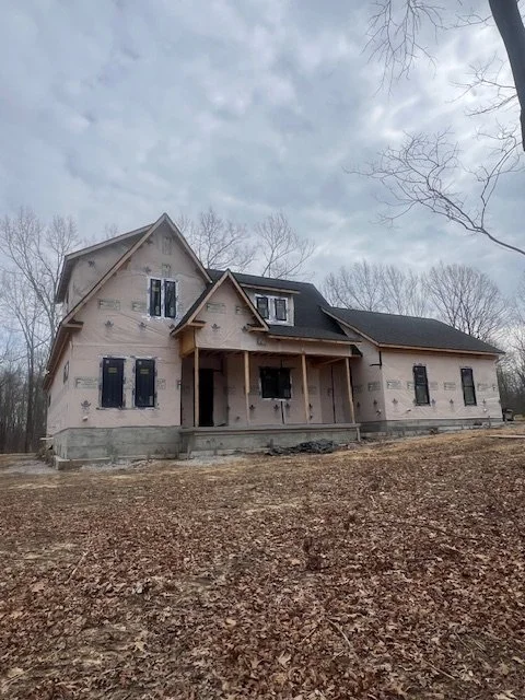 A two-story house under construction with a gabled roof and partially finished exterior, surrounded by leafless trees and dirt ground.