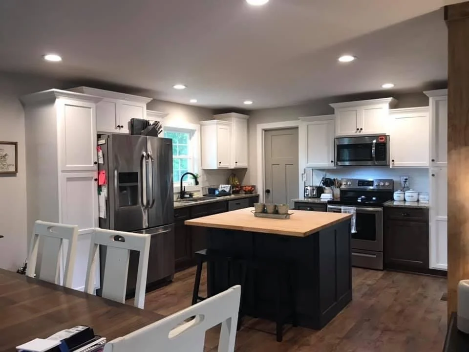 Modern kitchen with white upper cabinets, dark lower cabinets, stainless steel appliances including a refrigerator and stove, a kitchen island with a wooden top, and a dining table with white chairs. Remodeled Kitchen