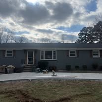 A gray single-story house with a large backyard, patio, and some outdoor furniture, under a partly cloudy sky.