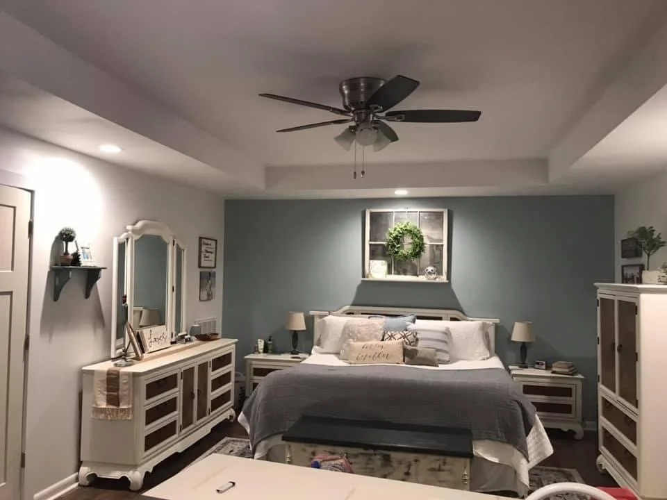 A cozy bedroom with a white bed, nightstands, and matching dressers, decorated with pillows, lamps, and plants, with a window and wall frame above the bed. Remodeled Bedroom