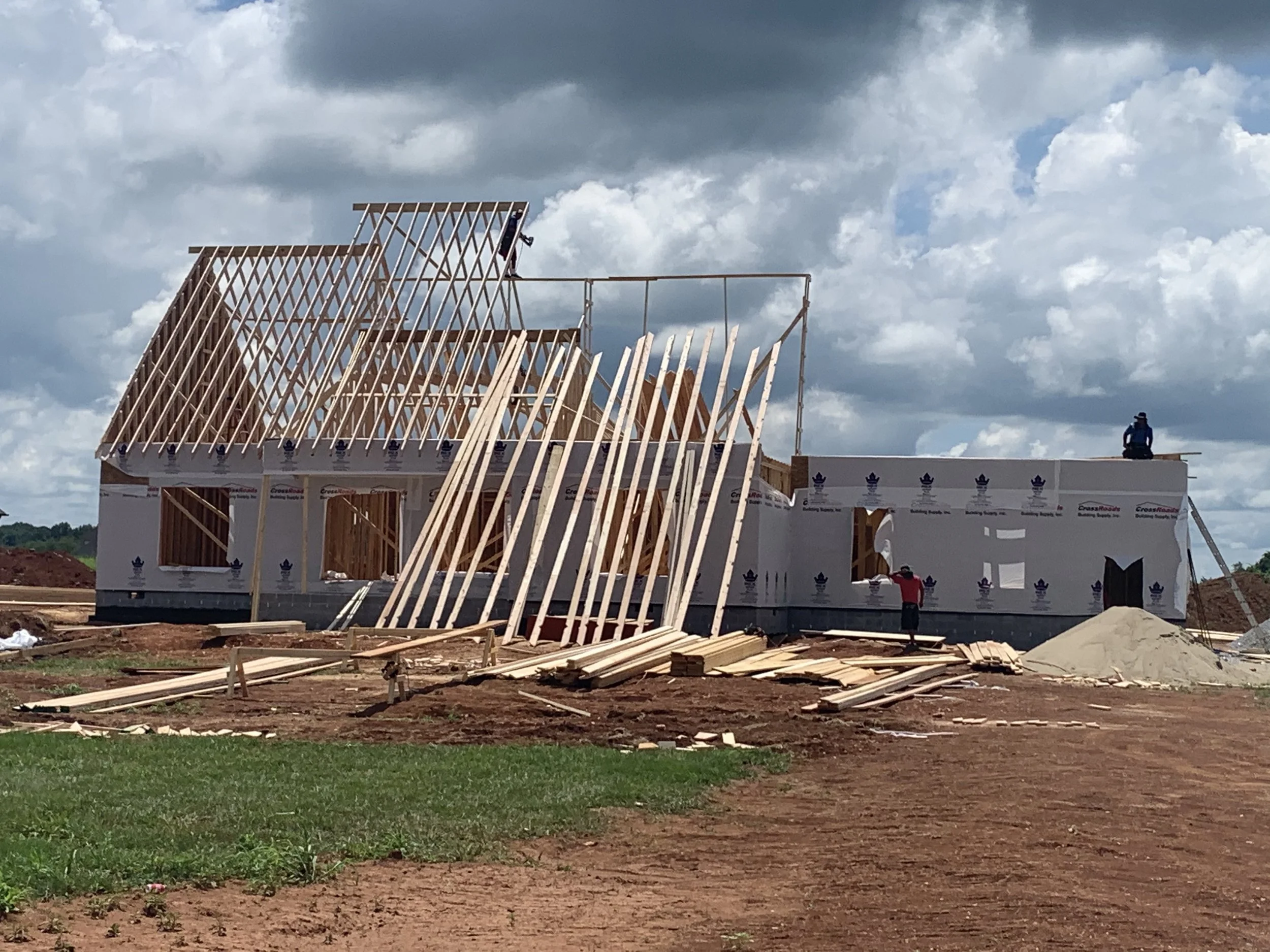 Construction site with a wooden house frame being built, and two workers on site under a cloudy sky. Building for a profit