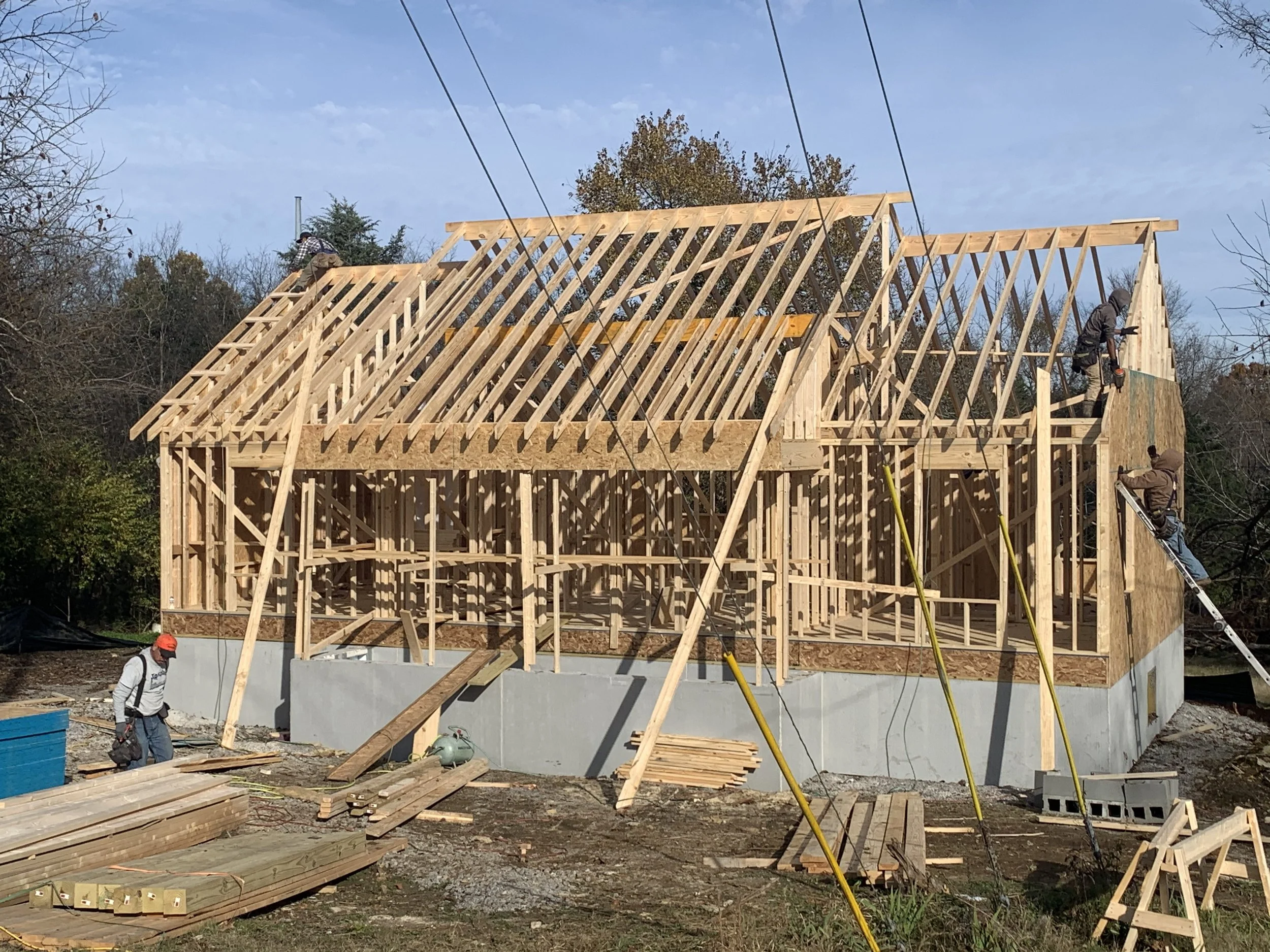 Wood framing construction of a house, with workers installing roof trusses, on a concrete foundation, in an outdoor setting with trees and a blue sky.