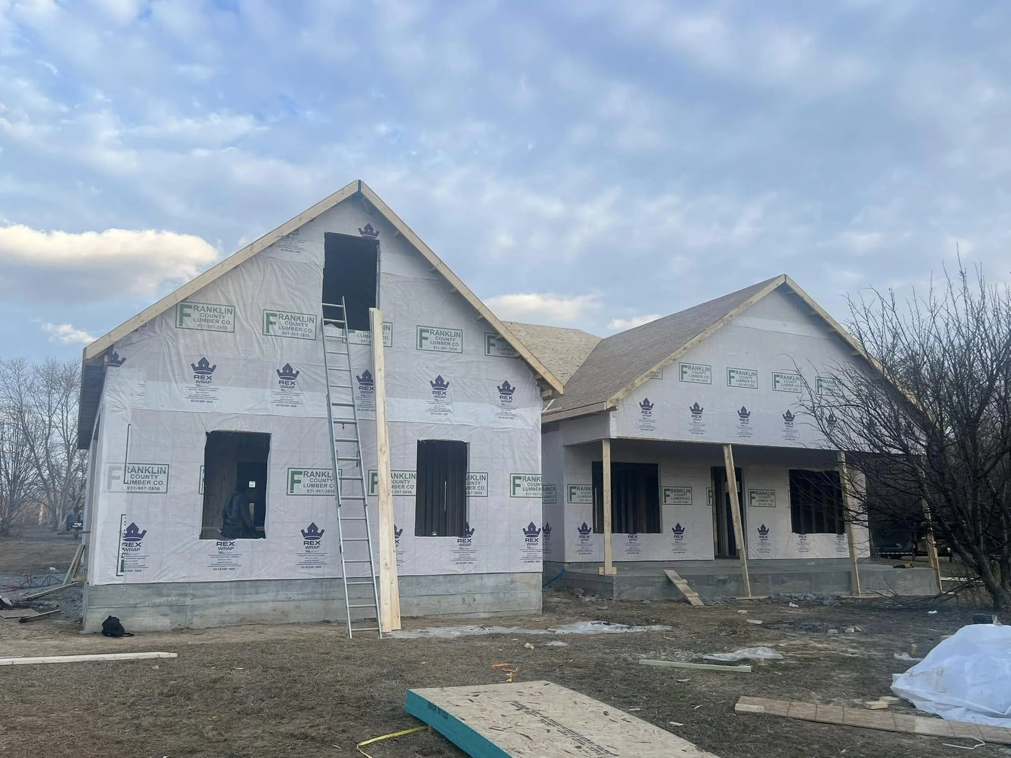 Under construction two-story house with exposed framing, wrapped in building paper, in a suburban area with cloudy sky and leafless trees.
