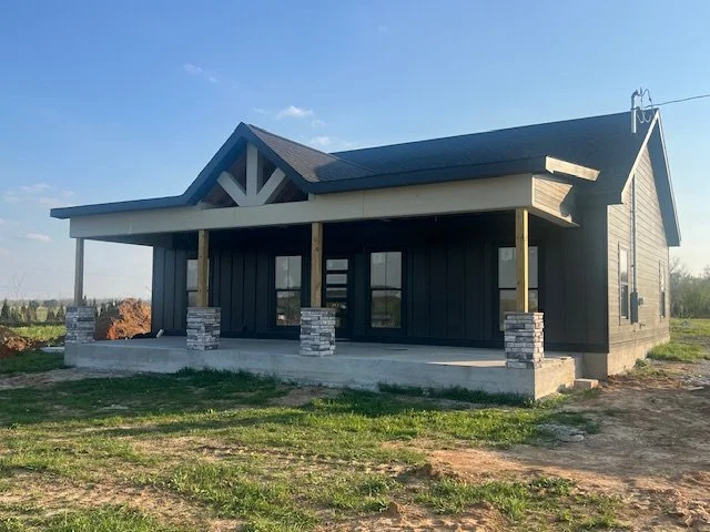 Newly built house with a front porch, black siding, and stone columns on a concrete foundation, under a clear sky.
