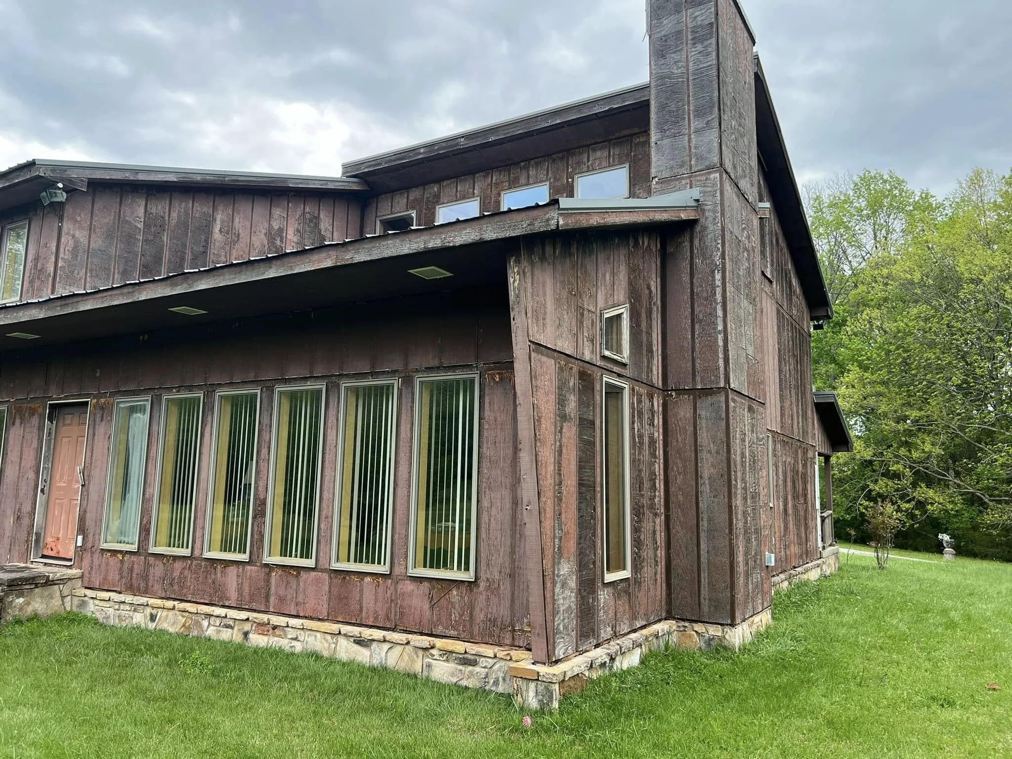A two-story wooden house with a stone foundation, brown vertical siding, multiple large windows with vertical blinds, and a door on the side, surrounded by green grass and trees under a cloudy sky.