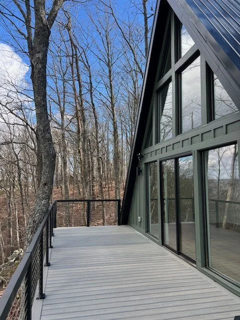 A balcony with a black railing attached to a modern house with large glass windows and a sloped metal roof, surrounded by leafless trees under a partly cloudy sky.