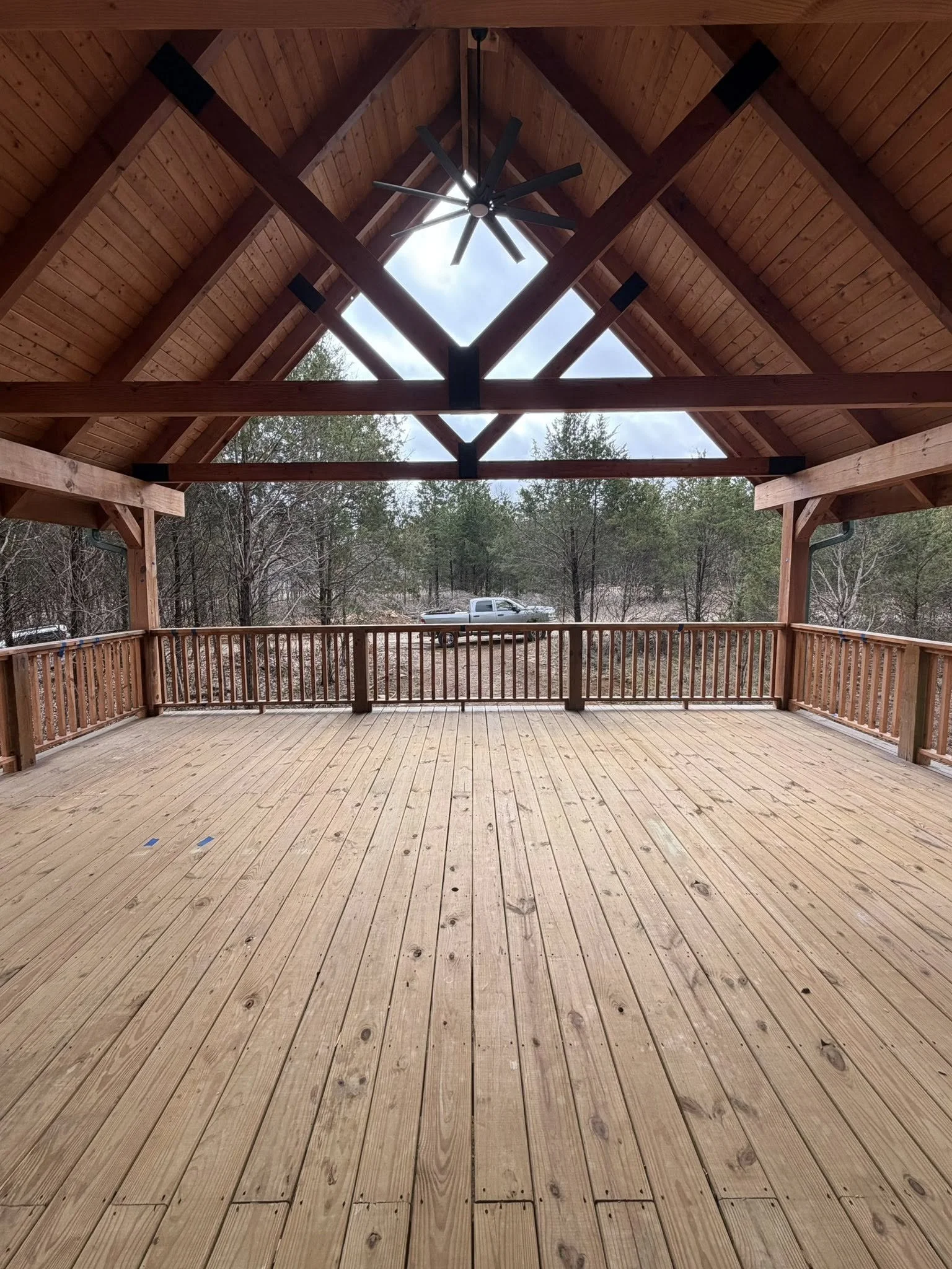 Empty wooden deck with railing, under a high, pointed wooden roof with a large black ceiling fan hanging from the center, overlooking trees and a vehicle in the background.