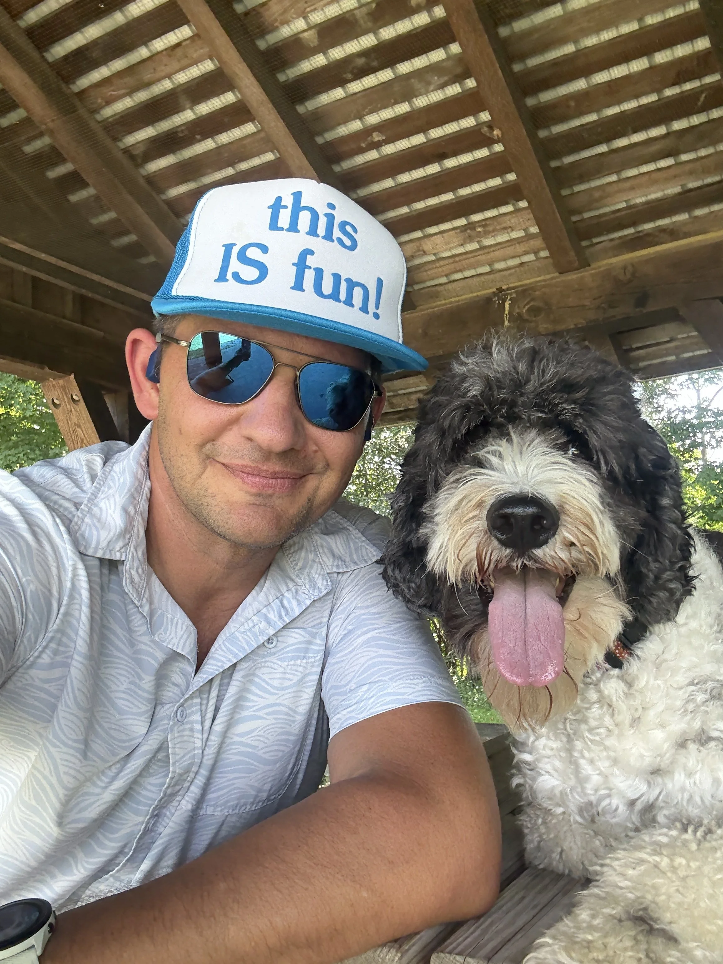 A man wearing a white patterned shirt, sunglasses, and a trucker hat with the words 'this IS fun!' taking a selfie with a happy black and white dog with its tongue out outdoors under a wooden shelter.