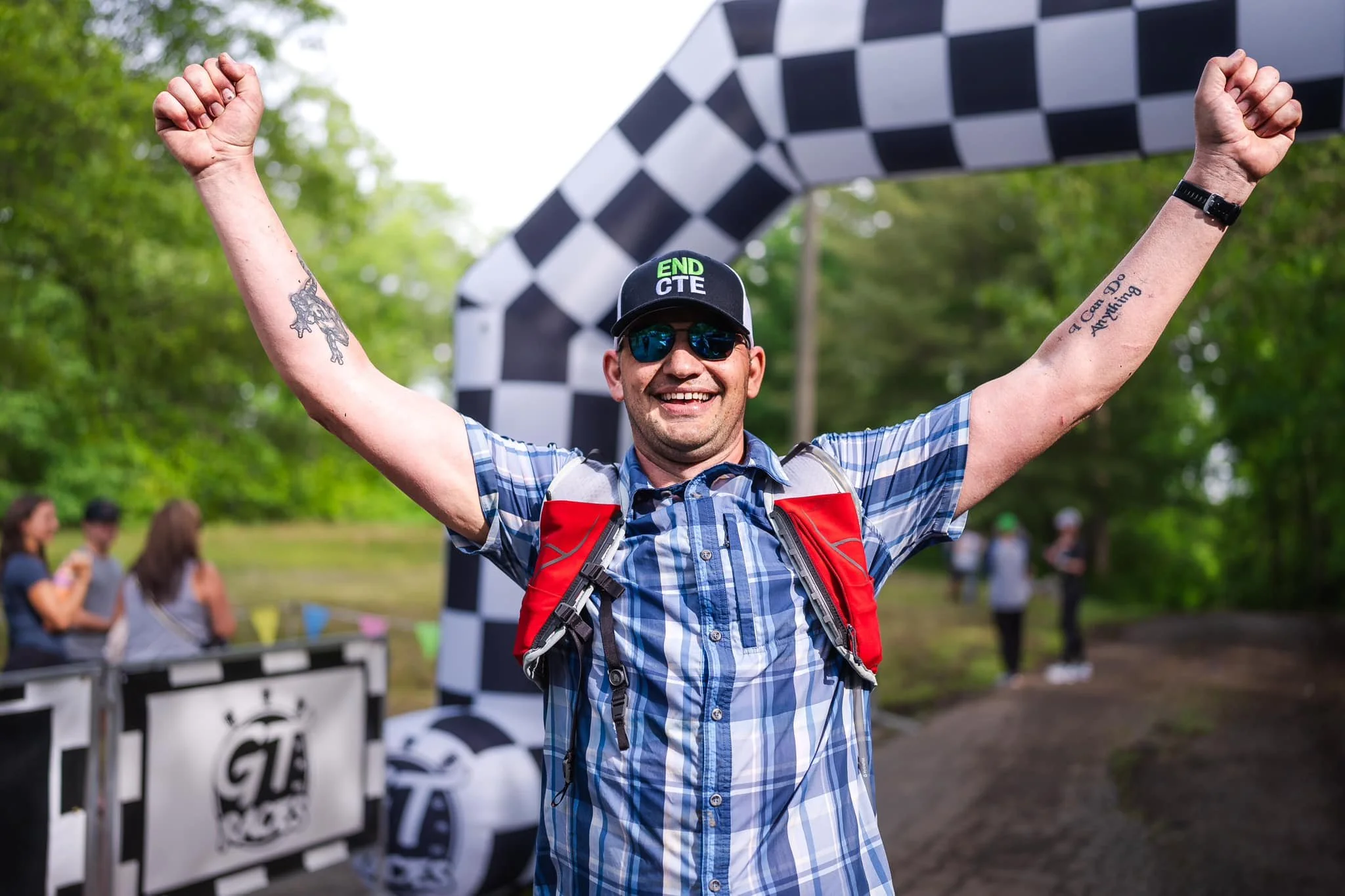 Man celebrating at a race finish line with arms raised, wearing sunglasses, a cap, and a backpack, in a wooded outdoor setting.