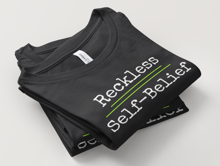 Stack of black t-shirts with white and green text reading 'Reckless self-belief' folded on a white surface.