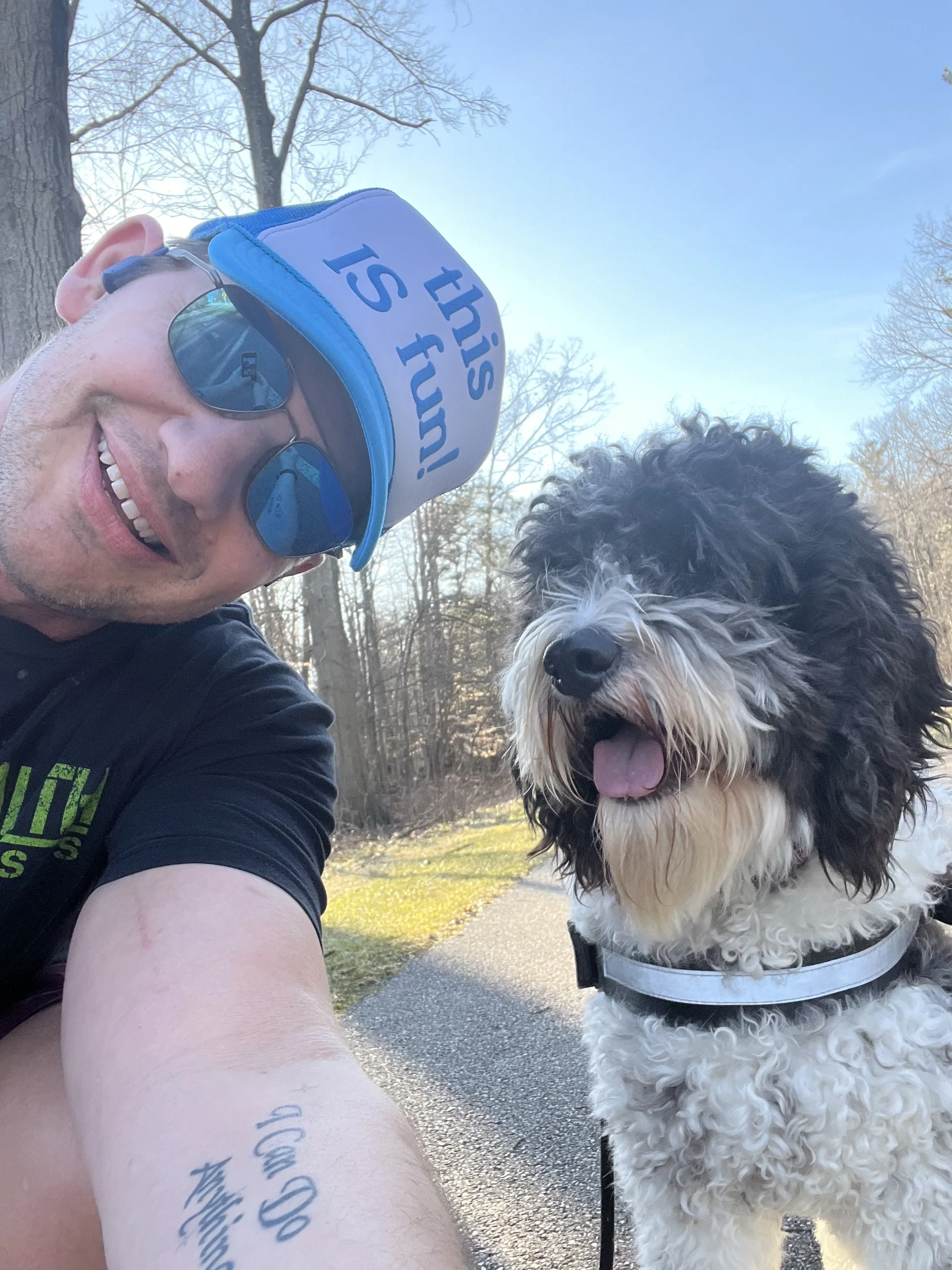 A man wearing sunglasses, a blue cap with the phrase "this is fun!", and a black t-shirt, smiling while taking a selfie with a fluffy black and white dog on a sunny walk in a park with bare trees.