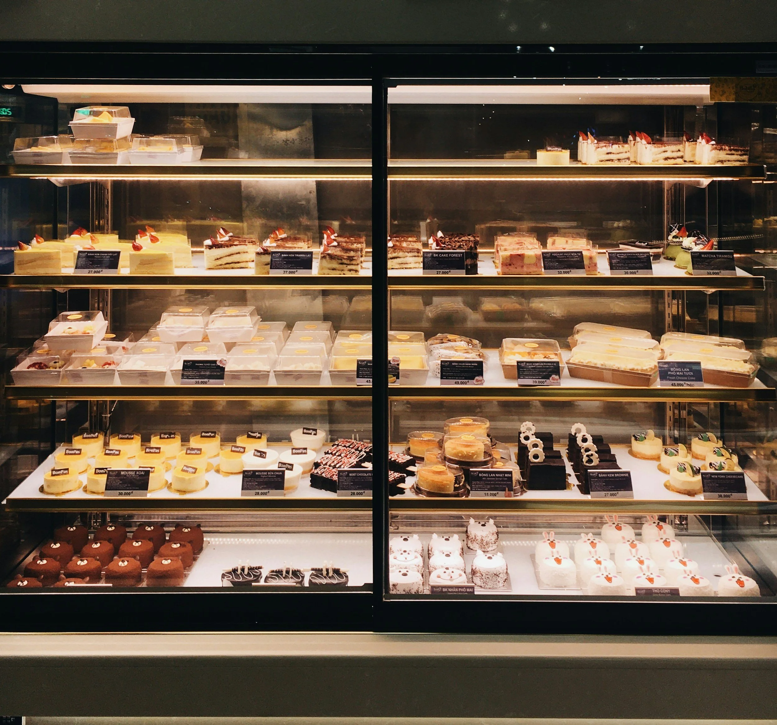 Display case filled with an assortment of cakes and desserts, including chocolate, strawberry, and fruit-topped cakes, neatly arranged on shelves in a bakery or cafe.