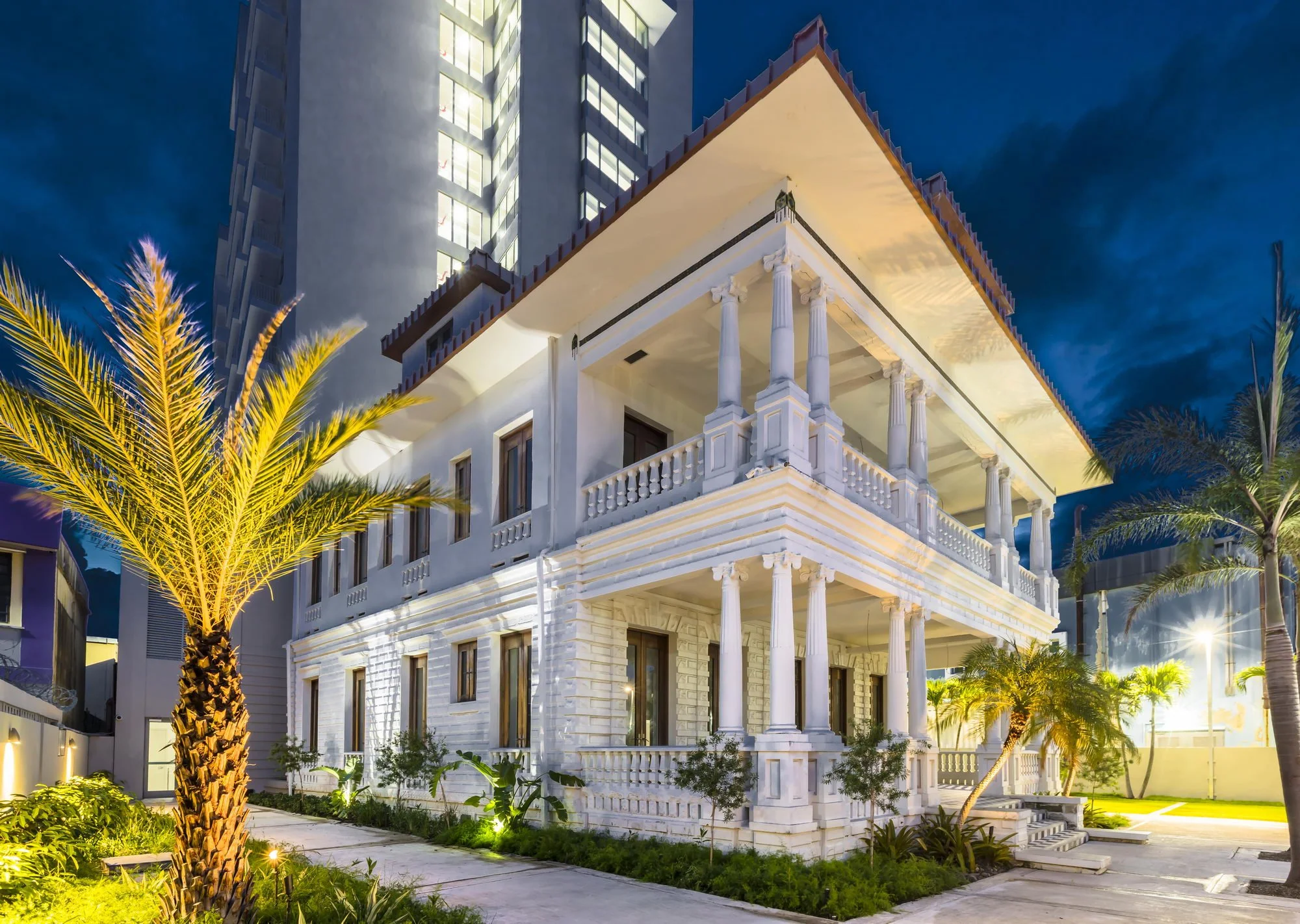 A large modern house with a traditional plantation-style porch, columns, and balcony railing, illuminated at night, with palm trees and garden plants in the foreground.