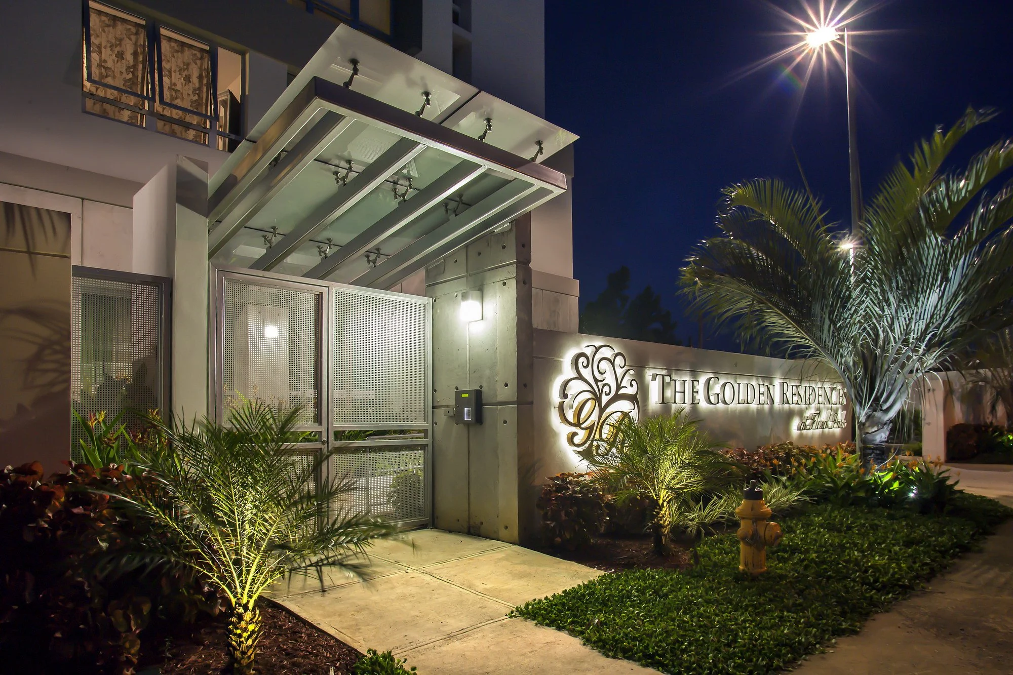 Night view of the entrance to The Golden Residence, a modern building with illuminated sign. The area is landscaped with plants and trees, and there is a prominent streetlamp shining brightly in the sky.