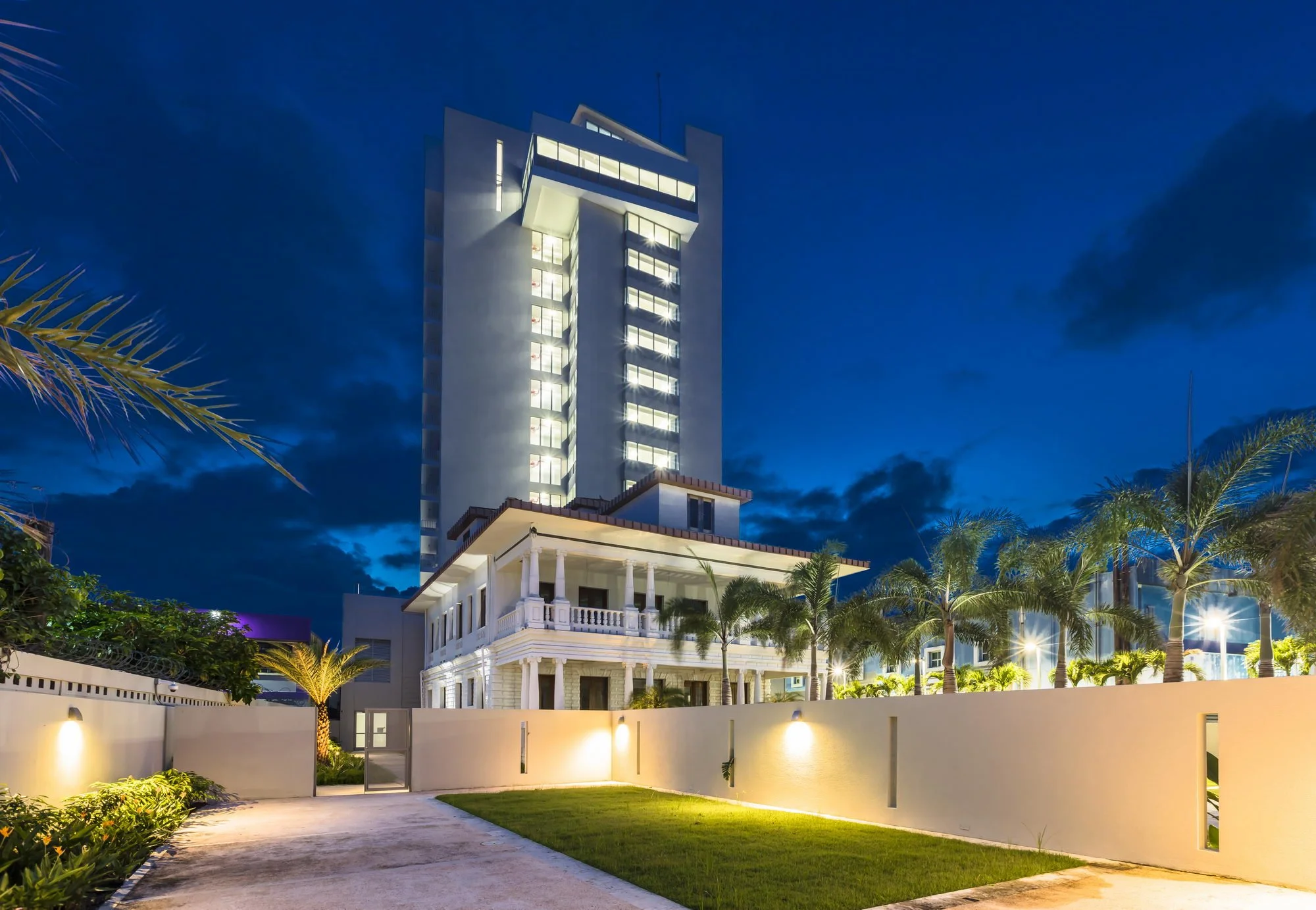Night view of a modern high-rise building illuminated against a dark sky, with palm trees and a white fence in the foreground.