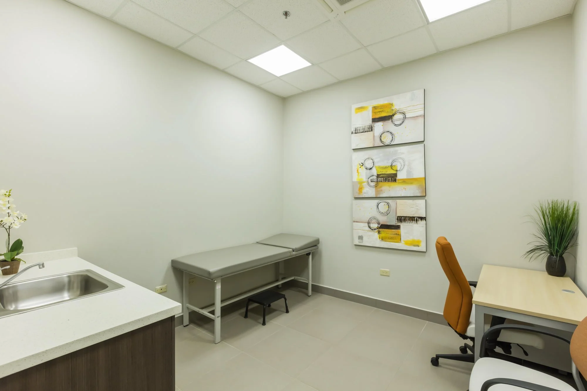 A medical examination room with white walls, a ceiling with fluorescent lights, and a painting with abstract designs in yellow, black, and white. There is a gray examination table, a small black stool, a desk with an orange chair, a plant in a black 