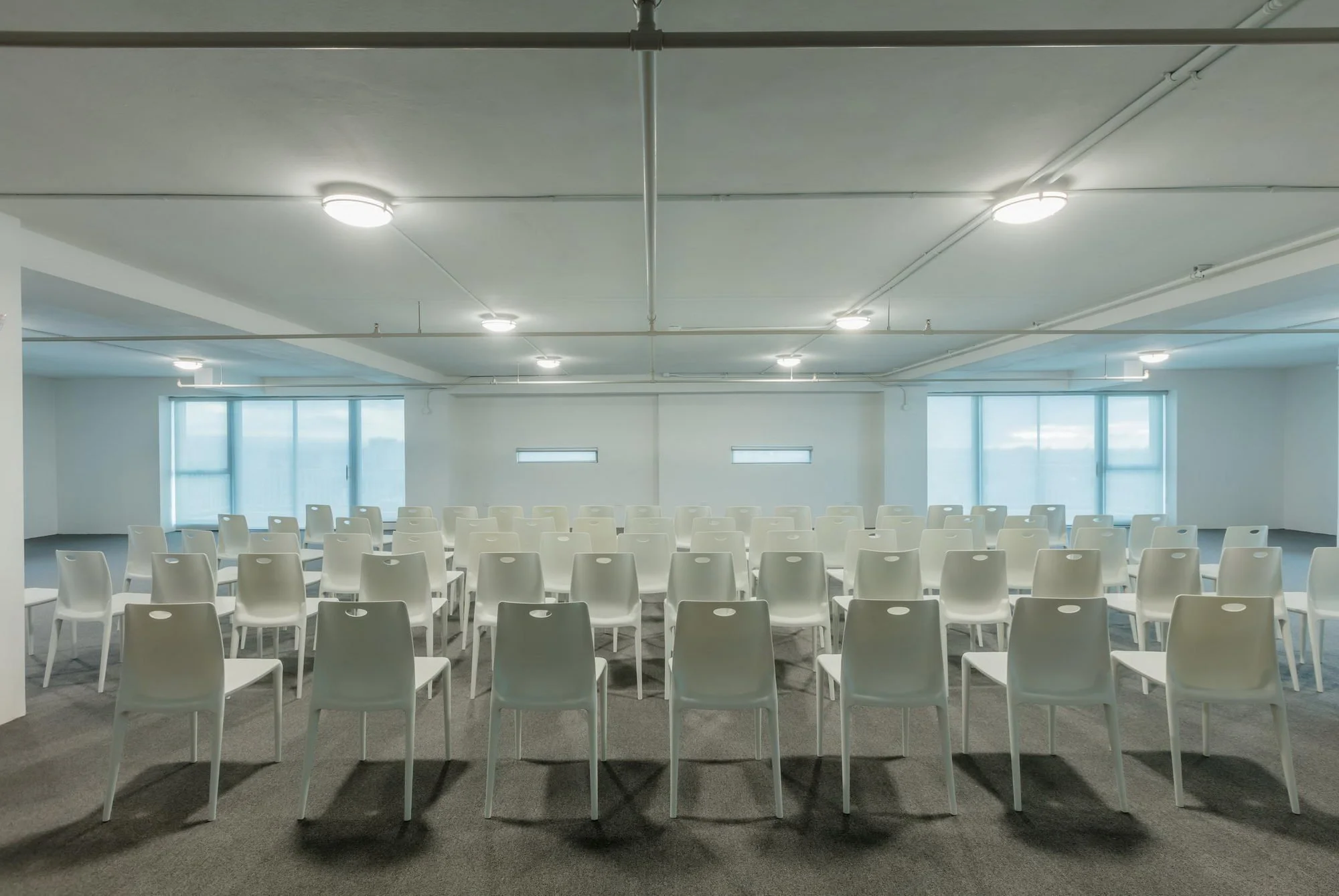 Empty conference or seminar room with rows of white chairs facing a front wall with large windows, ceiling lights, and a white ceiling