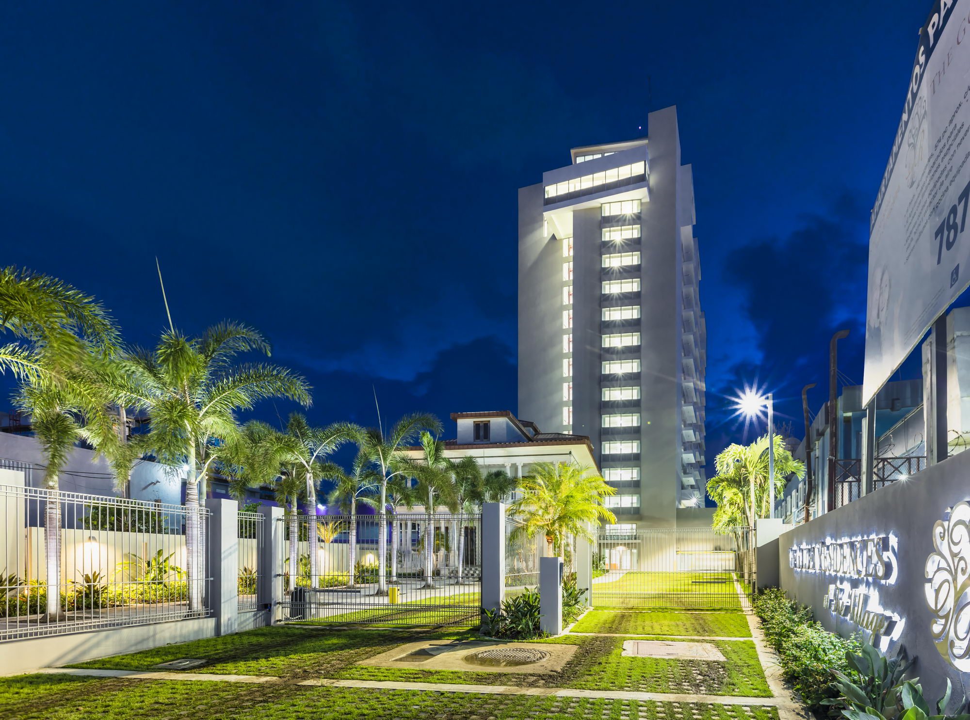Nighttime view of a tall, modern building with illuminated windows, surrounded by palm trees, a manicured lawn, and a white fence. A streetlamp shines in the foreground, with a sign partially visible on the right side.