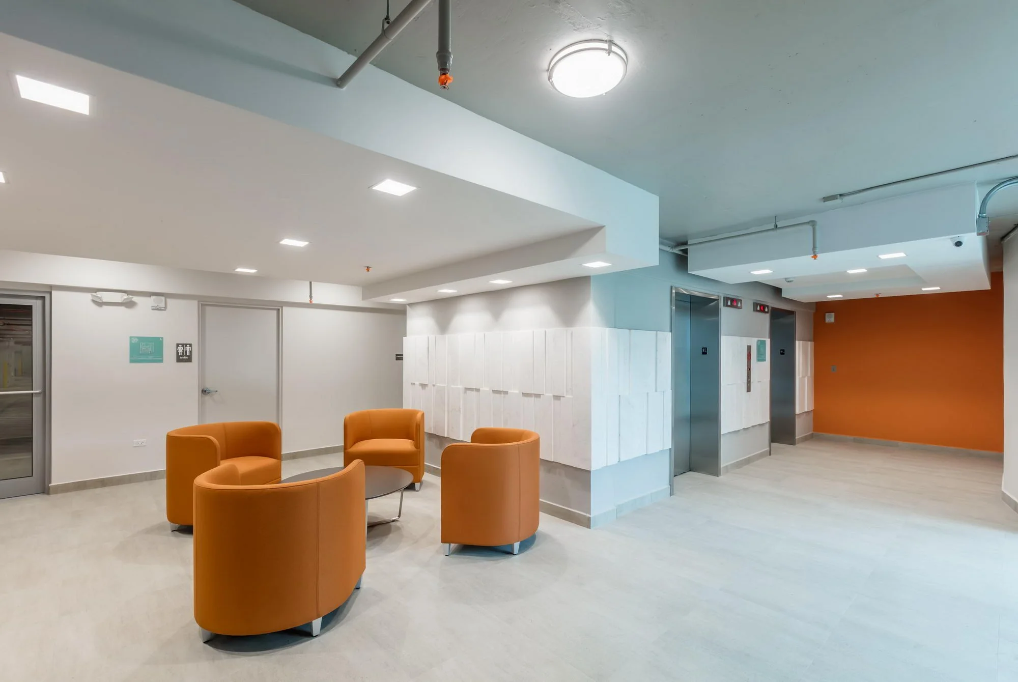 Interior view of a modern building lobby with orange chairs arranged in a circle, light-colored walls, ceiling lights, and an elevator with blue doors in the background, and two fire sprinklers on the ceiling.