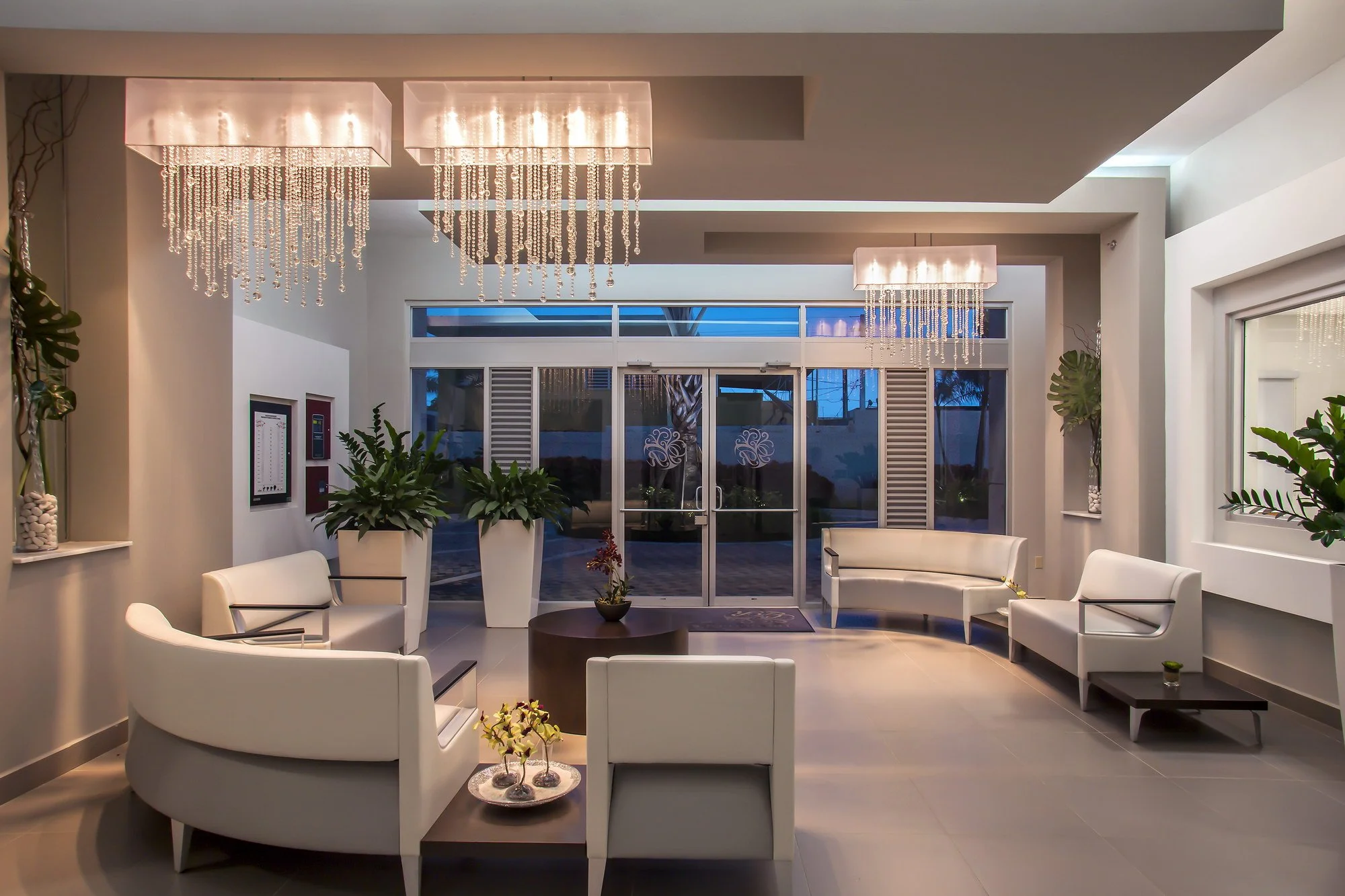 Modern lobby with white seating, large potted plants, a central round table with flowers, chandelier lights, and glass entrance doors with blue tint.