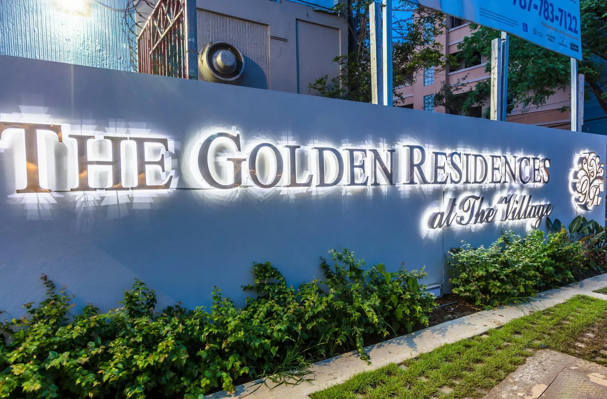 Sign at The Golden Residences at The Village, with greenery in front and apartment buildings in the background. The sign is illuminated and has decorative elements.