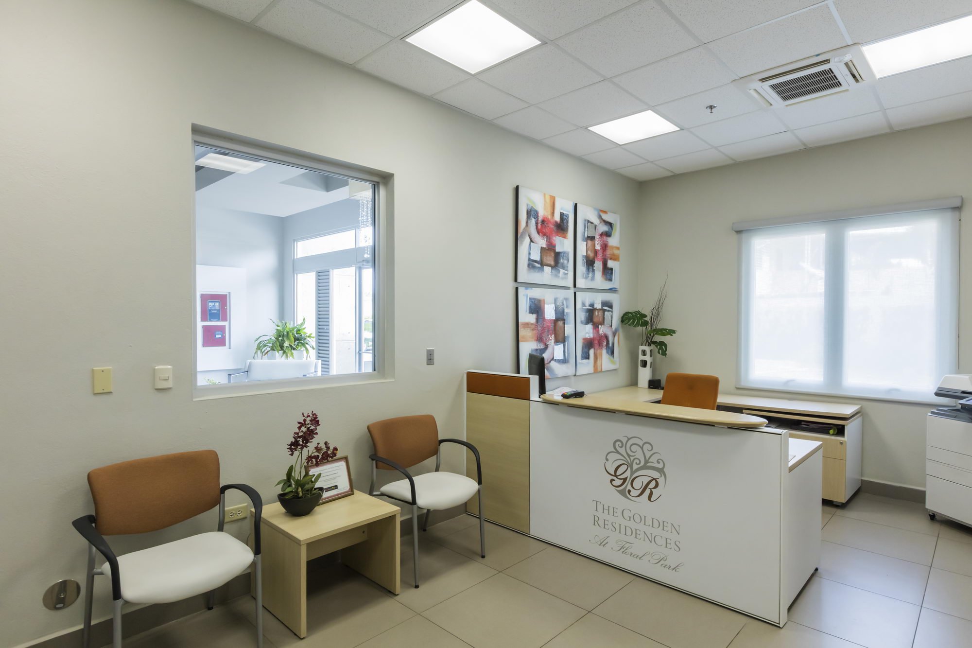 An interior view of a modern medical or office reception area with beige chairs, a small table with a plant, and a reception desk with decorative logo and text. There are abstract paintings on the wall, two windows, and ceiling lights.