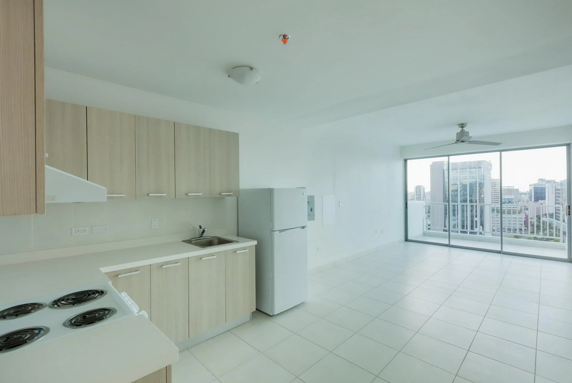 Modern kitchen with white countertops, light wood cabinets, a white refrigerator, stove, and a large balcony with city view through sliding glass doors.
