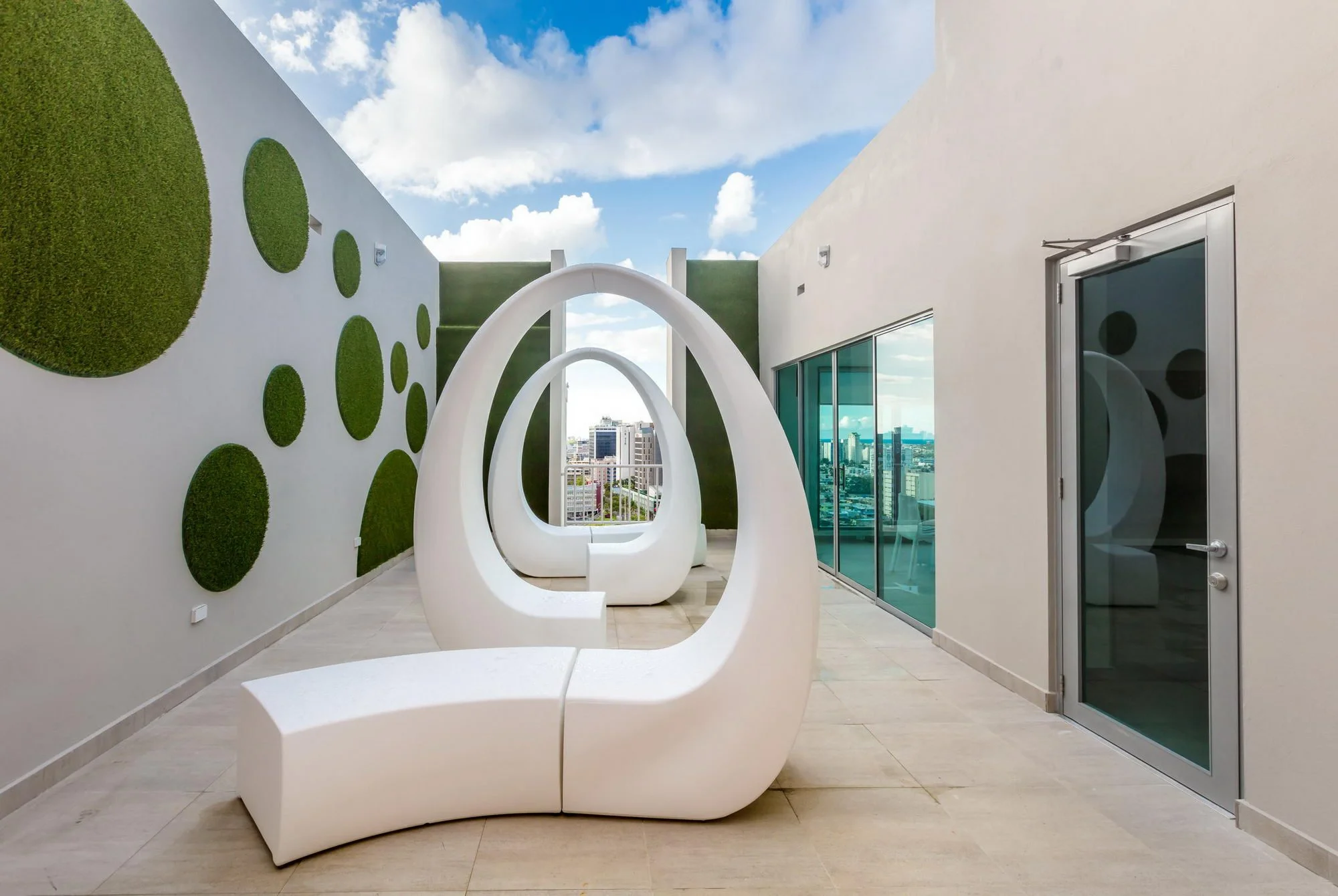 Modern rooftop terrace with white circular art installation, green wall accents, glass doors, and cityscape view under partly cloudy sky.