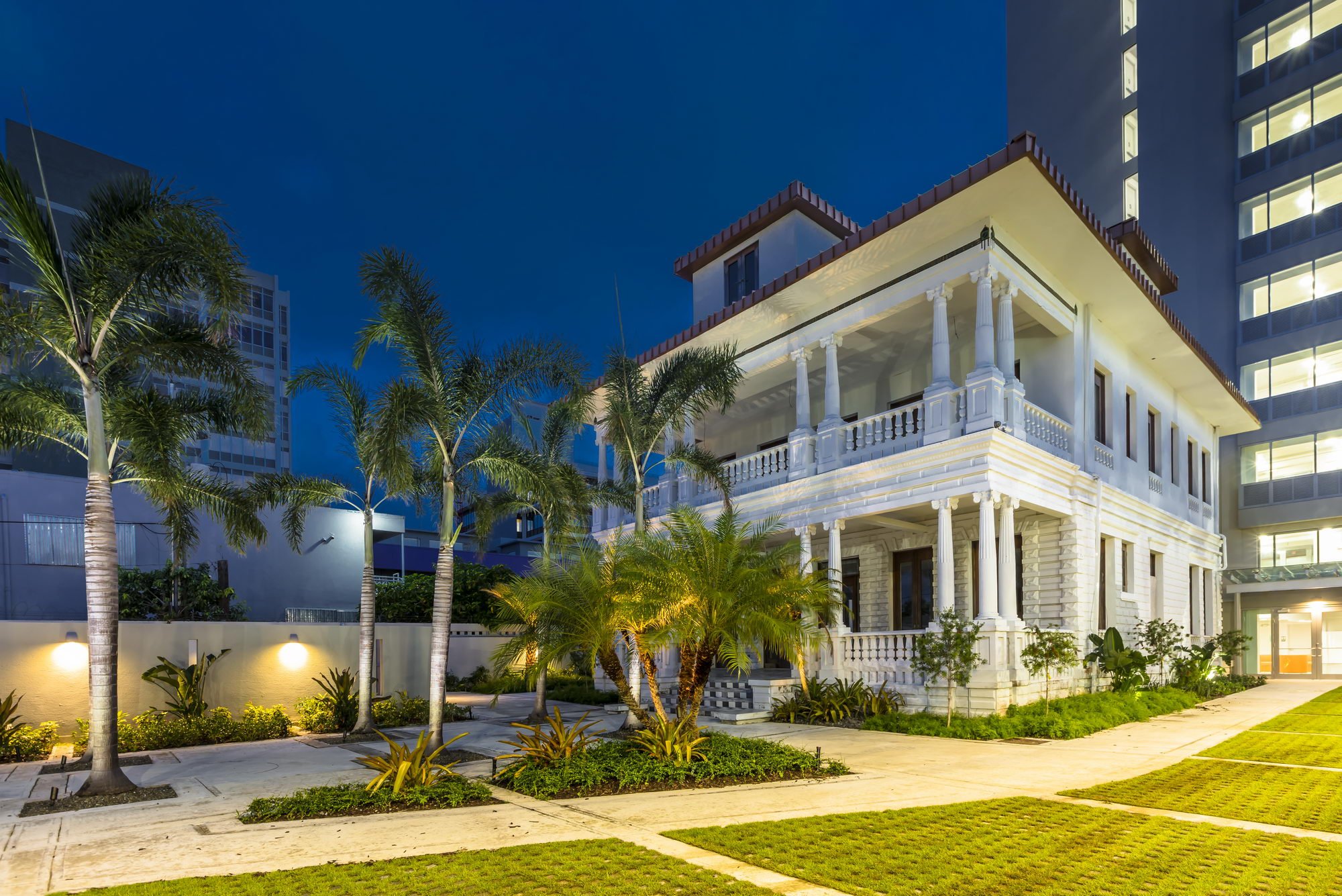 A multi-story house with classic architecture, white exterior, and tall columns in front, surrounded by a garden with palm trees and plants, at night with lights illuminating the scene.