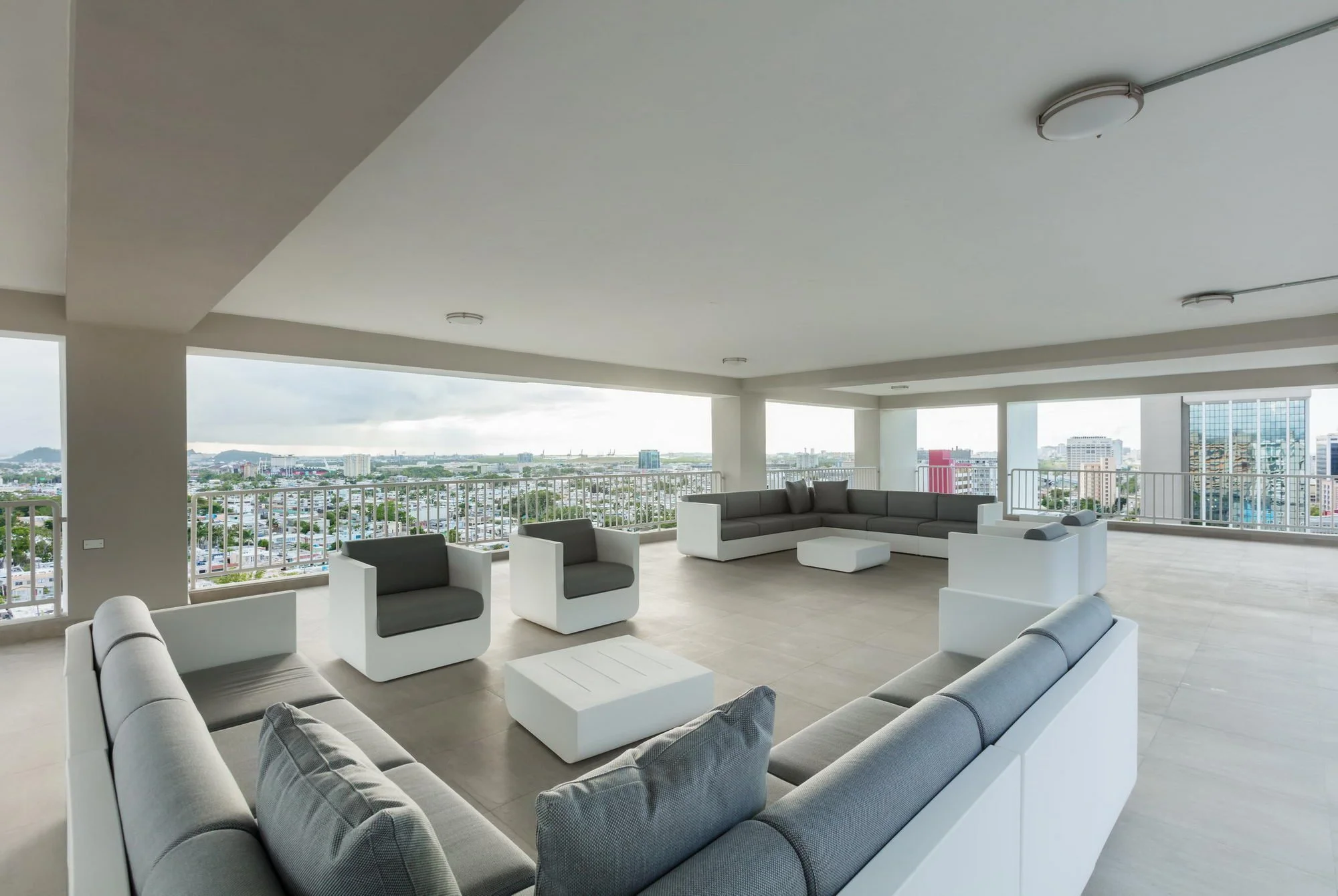 Modern outdoor rooftop lounge with white and gray furniture, overlooking a cityscape with high-rise buildings and a cloudy sky.