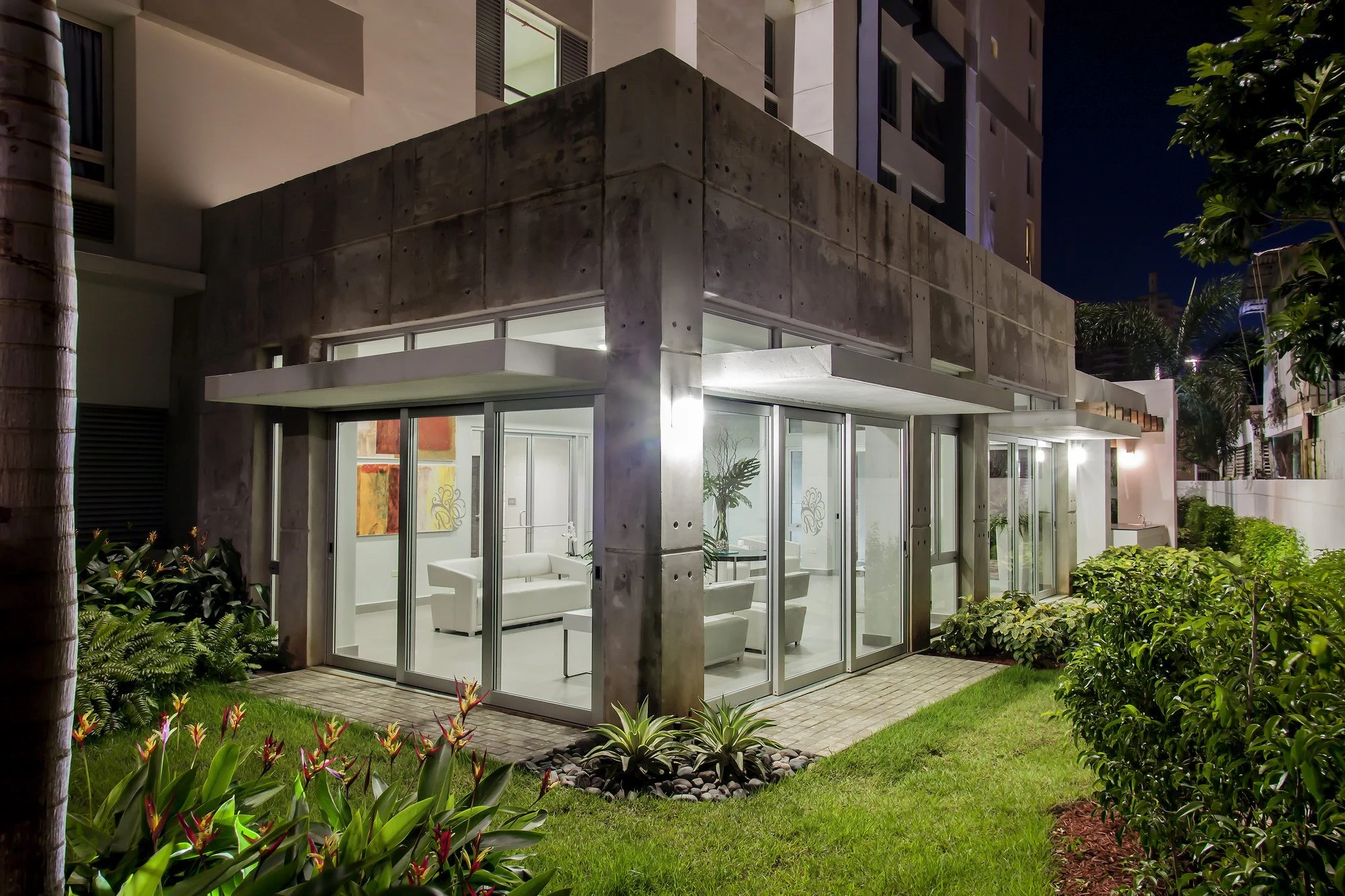 Exterior of a modern building at night with large glass windows, a patio, and lush landscaping including green plants and flowers.