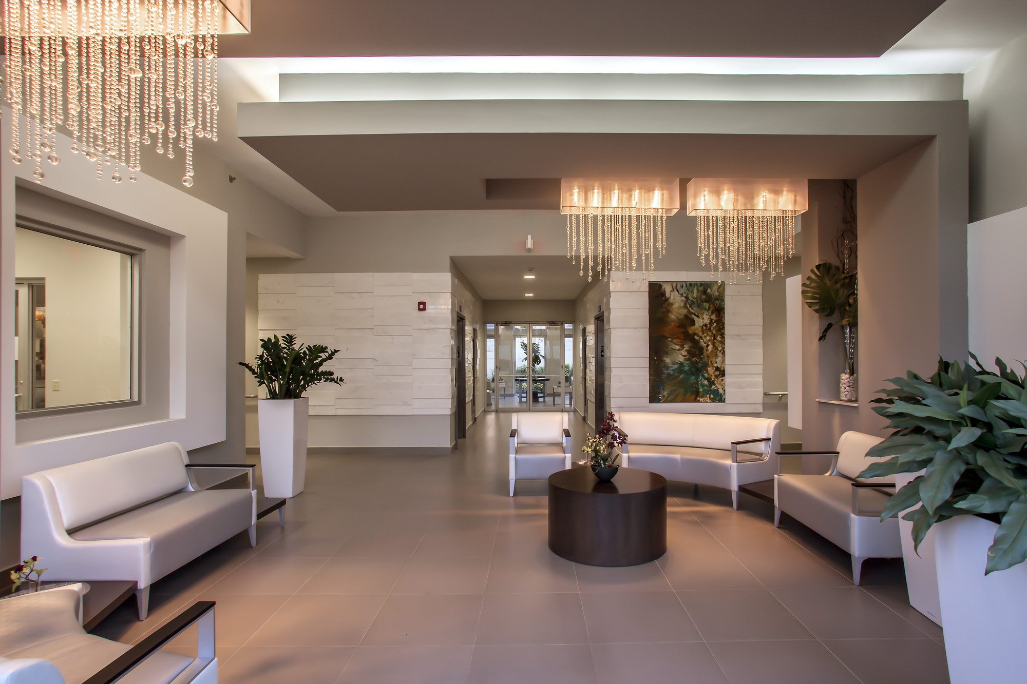 Modern hotel lobby with white furniture, indoor plants, abstract artwork, and contemporary lighting fixtures.