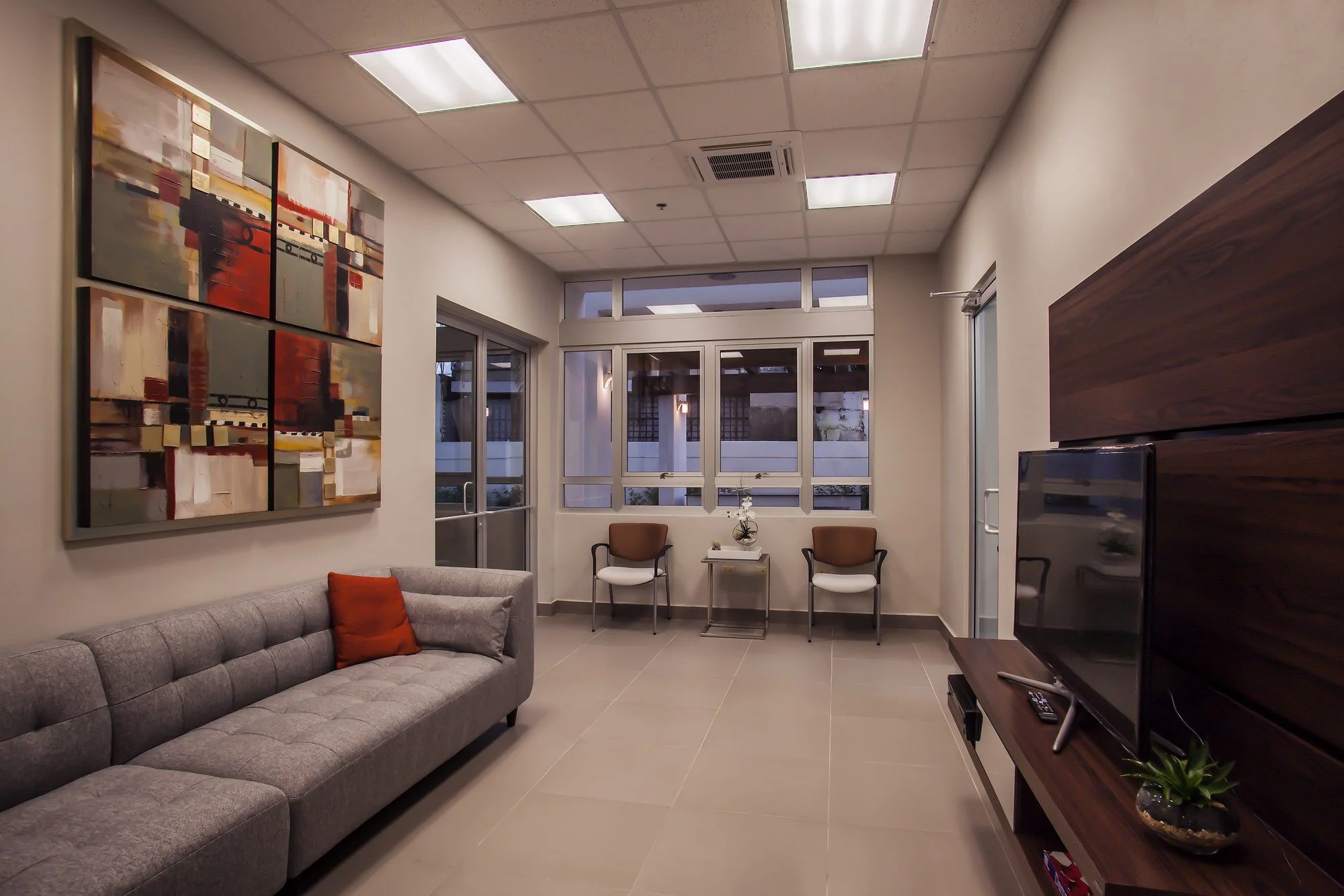 A waiting room with a gray tufted sofa with a red pillow, two brown chairs, a white side table with a potted orchid, large windowed doors, abstract wall art, and a flat-screen TV on a wooden stand in a well-lit space.