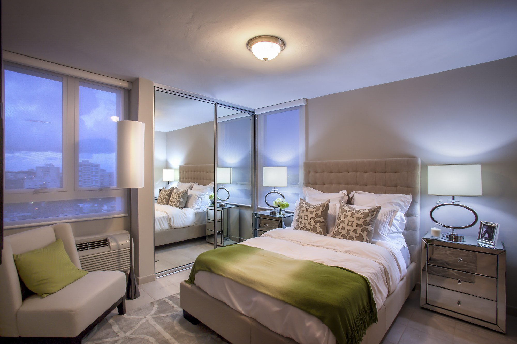A modern hotel room with two beds, each with white bedding and patterned pillows. There are nightstands with lamps, large windows with city views, and reflective closet doors. The room has neutral tones with green accents on the beds and a seating ar