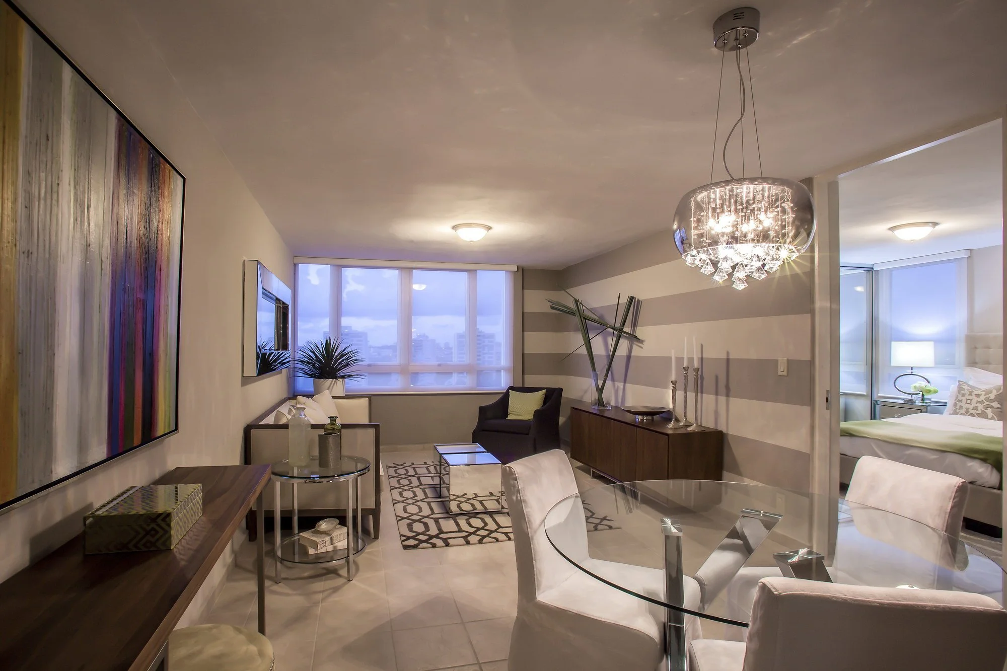 Interior of a modern apartment living room with a chandelier, large window showing cityscape, and various furniture including a glass dining table, white chairs, and a dark armchair.