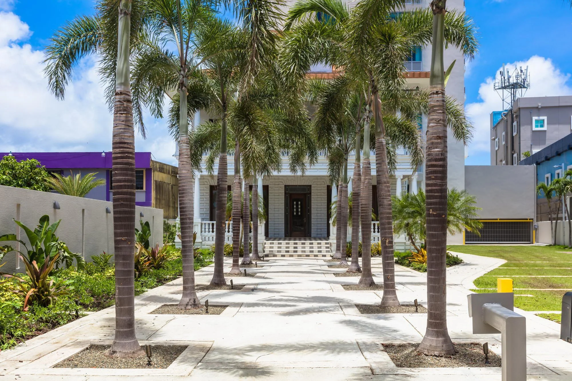 A modern residential building with a white facade, flanked by tall palm trees and landscaped greenery, under a bright blue sky with some clouds.