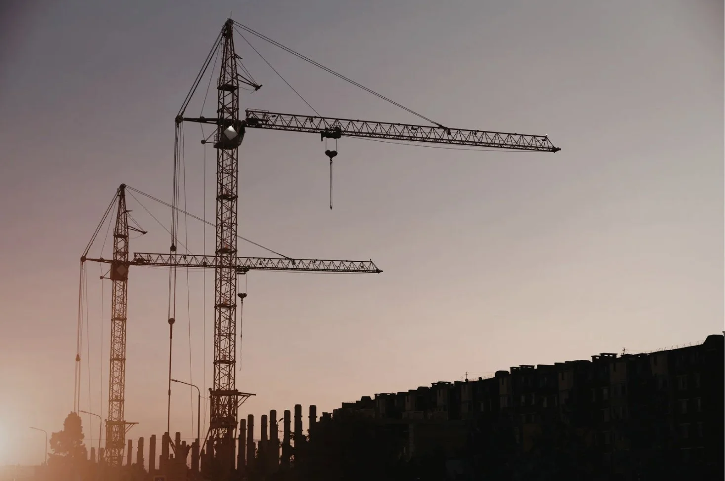 Silhouettes of two construction cranes against a dusk sky near a construction site with a partially built wall.