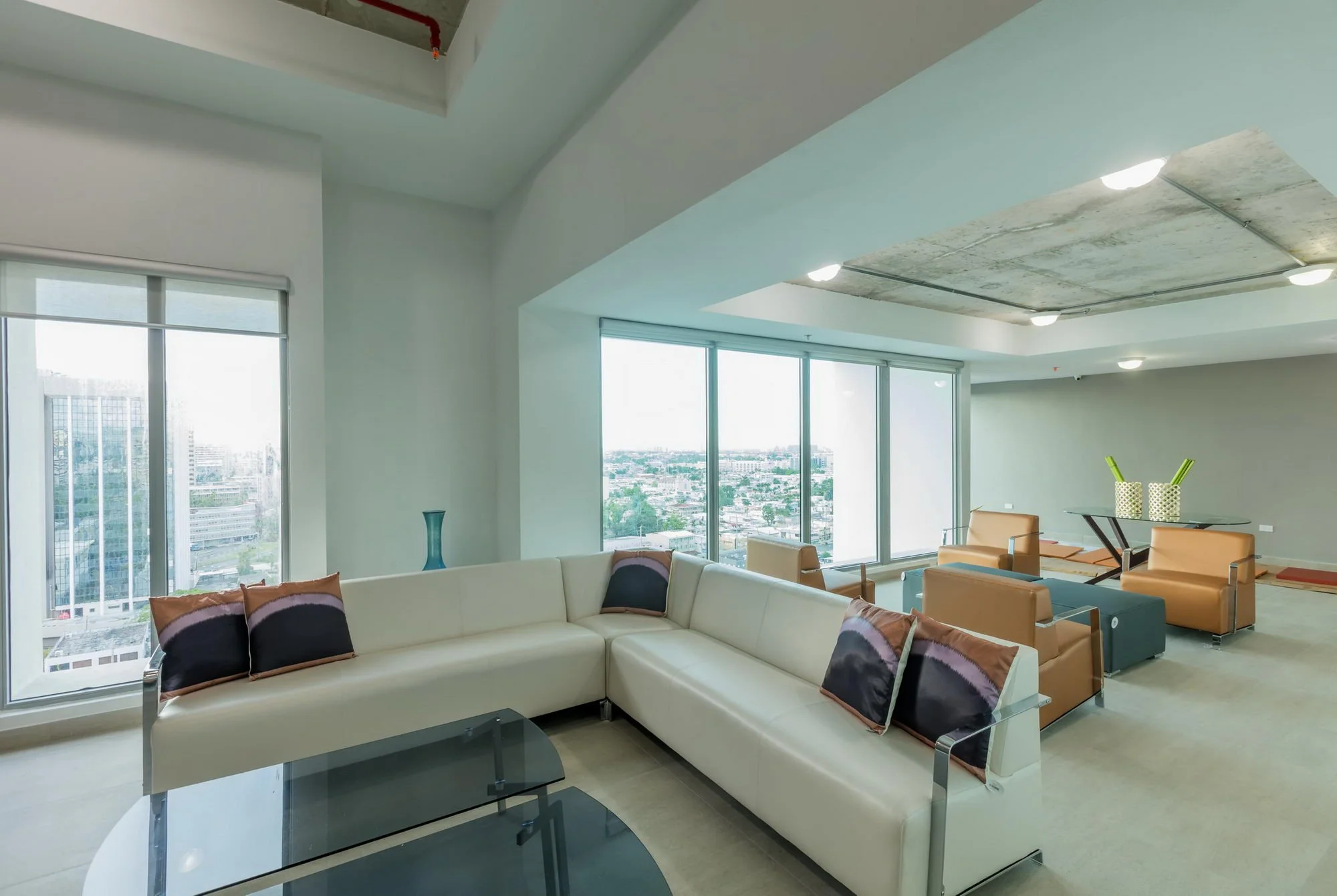 Modern living room with white sectional sofa, decorative pillows, glass coffee table, tan chairs, large windows showing cityscape, ceiling with exposed concrete section, recessed lighting.