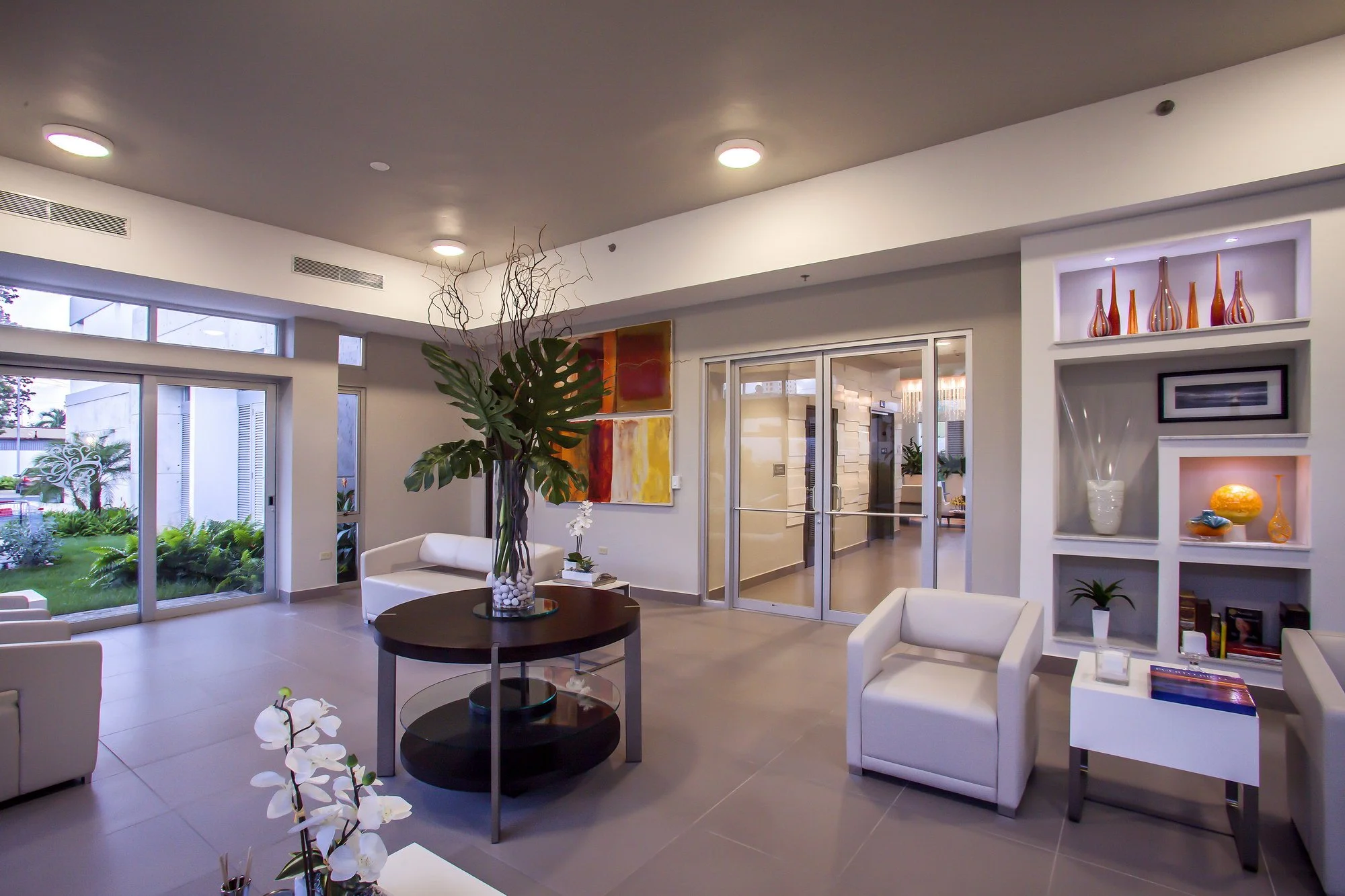 Modern interior living room with white furniture, large window, green plants, colorful artwork, and decorative vases on shelves.