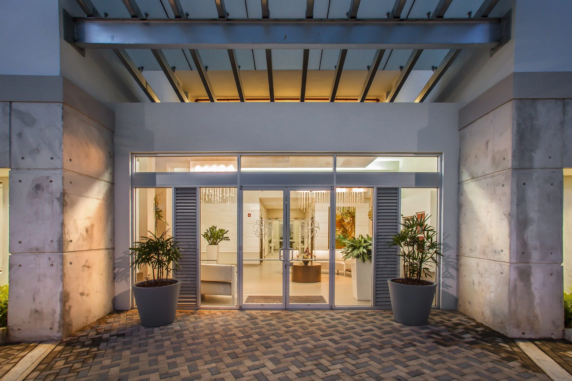 Exterior view of a modern building entrance with glass doors, potted plants, and a brick pathway, taken during evening.