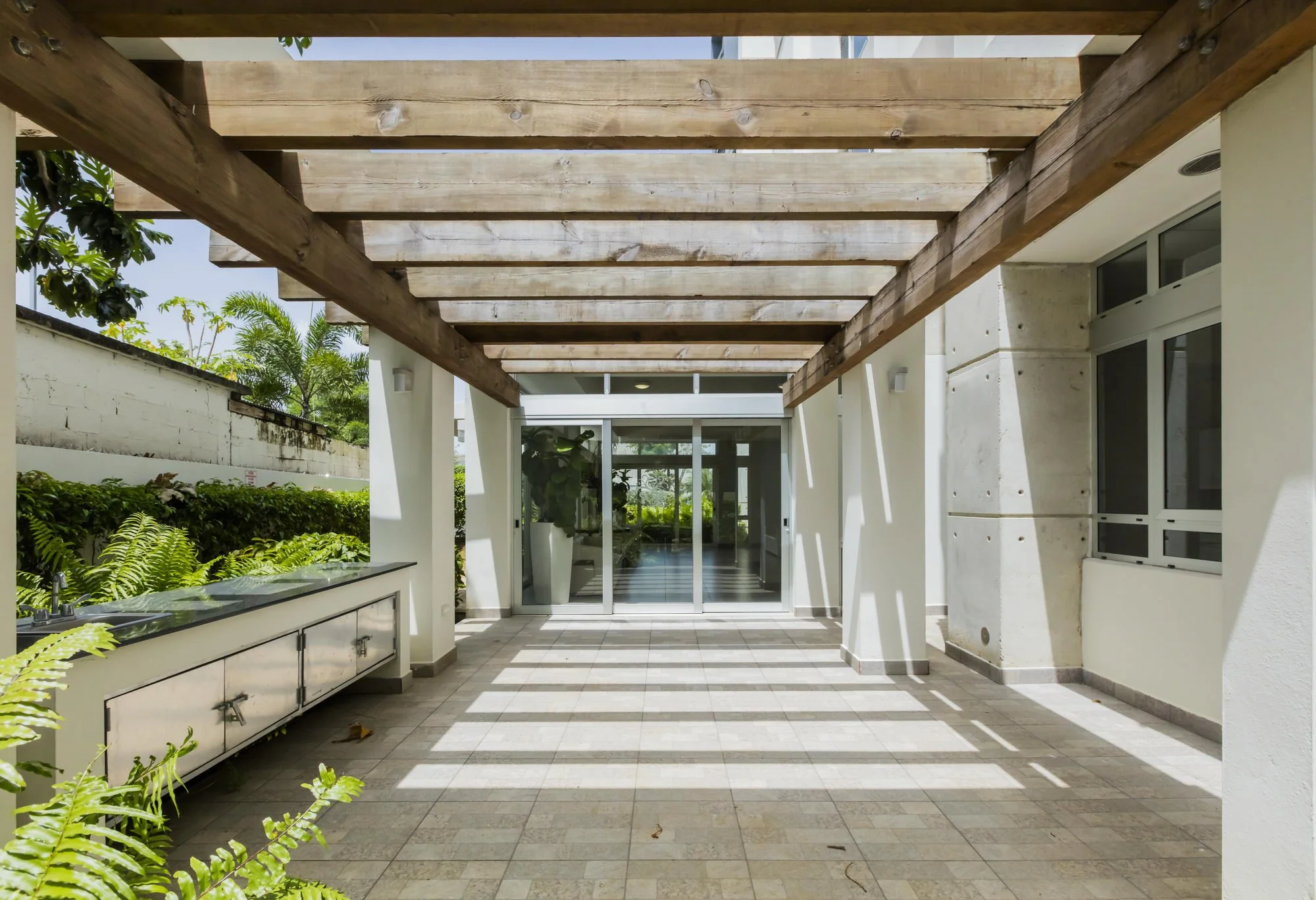 A covered outdoor patio area with a wooden pergola overhead, a sink with cabinets to the left, and a glass door leading inside a building. There are lush green plants nearby and sunlight casting shadows on the tiled floor.