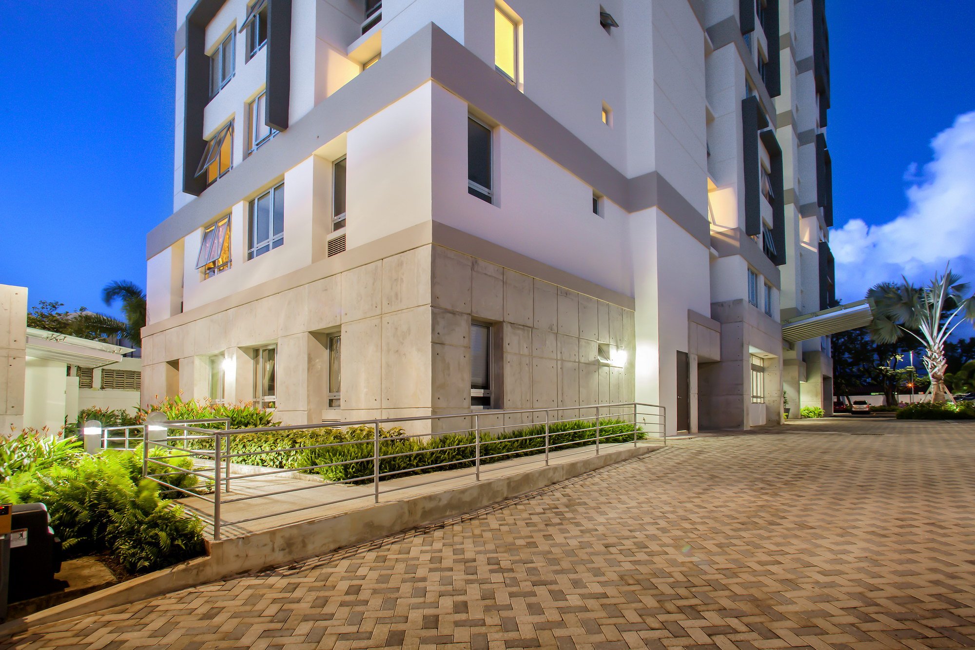 Exterior view of a modern white apartment building with several windows, some open, illuminated at dusk. The ground level features a paved walkway, landscaped greenery, and exterior lighting.
